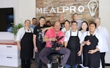 A group of ten people are standing and smiling in front of a brick wall with the word 'MEALPRO' displayed along with a logo of a light bulb and utensils. The group consists of both men and women, some wearing aprons and chef coats, suggesting a culinary setting. The man in the center is seated and holding a professional camera, indicating a photo or video shoot environment. There is a plant on a countertop to the left and a television screen partially visible.