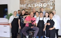 A group of ten people are standing and smiling in front of a brick wall with the word 'MEALPRO' displayed along with a logo of a light bulb and utensils. The group consists of both men and women, some wearing aprons and chef coats, suggesting a culinary setting. The man in the center is seated and holding a professional camera, indicating a photo or video shoot environment. There is a plant on a countertop to the left and a television screen partially visible.