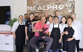A group of ten people are standing and smiling in front of a brick wall with the word 'MEALPRO' displayed along with a logo of a light bulb and utensils. The group consists of both men and women, some wearing aprons and chef coats, suggesting a culinary setting. The man in the center is seated and holding a professional camera, indicating a photo or video shoot environment. There is a plant on a countertop to the left and a television screen partially visible.