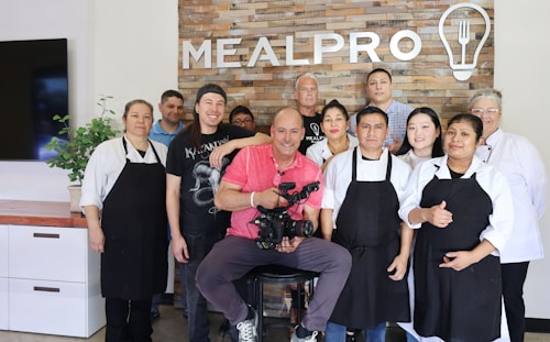 A group of ten people are standing and smiling in front of a brick wall with the word 'MEALPRO' displayed along with a logo of a light bulb and utensils. The group consists of both men and women, some wearing aprons and chef coats, suggesting a culinary setting. The man in the center is seated and holding a professional camera, indicating a photo or video shoot environment. There is a plant on a countertop to the left and a television screen partially visible.