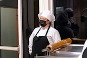A person wearing a white chef's uniform, including a chef hat and black apron, stands in a kitchen or food preparation area. The individual is also wearing a black face mask. Behind them is a reflective glass surface, showing their reflection. A piece of kitchen equipment with a cylindrical, amber-colored component is visible in the foreground.