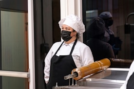 A person wearing a white chef's uniform, including a chef hat and black apron, stands in a kitchen or food preparation area. The individual is also wearing a black face mask. Behind them is a reflective glass surface, showing their reflection. A piece of kitchen equipment with a cylindrical, amber-colored component is visible in the foreground.