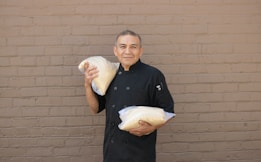 A person in a black chef's coat is holding two large bags of white grains against a beige brick wall.