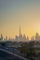 A cityscape of Dubai at sunset with the skyline prominently featuring the Burj Khalifa. The sky is filled with warm hues as the sun sets behind the iconic buildings. Palm trees and a road with moving cars are visible in the foreground, framed by the sprawling urban landscape.