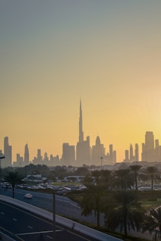 A cityscape of Dubai at sunset with the skyline prominently featuring the Burj Khalifa. The sky is filled with warm hues as the sun sets behind the iconic buildings. Palm trees and a road with moving cars are visible in the foreground, framed by the sprawling urban landscape.