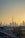 A cityscape of Dubai at sunset with the skyline prominently featuring the Burj Khalifa. The sky is filled with warm hues as the sun sets behind the iconic buildings. Palm trees and a road with moving cars are visible in the foreground, framed by the sprawling urban landscape.