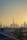 A cityscape of Dubai at sunset with the skyline prominently featuring the Burj Khalifa. The sky is filled with warm hues as the sun sets behind the iconic buildings. Palm trees and a road with moving cars are visible in the foreground, framed by the sprawling urban landscape.