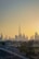 A cityscape of Dubai at sunset with the skyline prominently featuring the Burj Khalifa. The sky is filled with warm hues as the sun sets behind the iconic buildings. Palm trees and a road with moving cars are visible in the foreground, framed by the sprawling urban landscape.