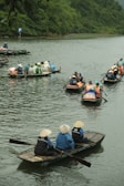 A group of people wearing traditional conical hats are seated in small wooden boats on a calm river, surrounded by lush green vegetation. Several boats are visible, each carrying multiple passengers, with some dressed in brightly colored raincoats. The scene conveys a sense of outdoor activity and cultural experience.