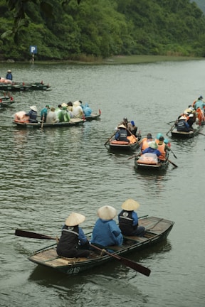 A group of people wearing traditional conical hats are seated in small wooden boats on a calm river, surrounded by lush green vegetation. Several boats are visible, each carrying multiple passengers, with some dressed in brightly colored raincoats. The scene conveys a sense of outdoor activity and cultural experience.