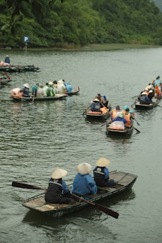A group of people wearing traditional conical hats are seated in small wooden boats on a calm river, surrounded by lush green vegetation. Several boats are visible, each carrying multiple passengers, with some dressed in brightly colored raincoats. The scene conveys a sense of outdoor activity and cultural experience.