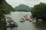 Several boats filled with people wearing life jackets are navigating along a river flanked by lush green trees and mountains. Some boats are lined up along a dock area on the left side of the image, while others are moving along the river.