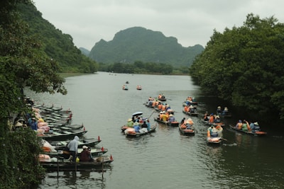 Several boats filled with people wearing life jackets are navigating along a river flanked by lush green trees and mountains. Some boats are lined up along a dock area on the left side of the image, while others are moving along the river.