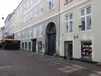 A street with buildings showcasing large windows and an arched doorway. A small bookshop named 'Soul Books' is visible with books displayed in the window. A restaurant with an outdoor seating area covered in an awning is situated further along the street. Cobblestone pavement adds a historical touch, and there are bicycles parked along the sidewalk.