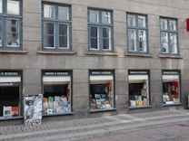 A row of windows on an urban building with a small bookstore at the ground level. The shop displays a variety of books and promotional material in the windows under the sign 'B&oslash;ger & Kuriosa'. Above the shop, there are several identical windows on a gray facade.