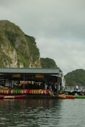 A floating structure houses numerous colorful kayaks lined up neatly in rows against a backdrop of lush green cliffs. The reflection of the kayaks and the structure is visible in the calm water. People are seen near the kayaks, suggesting an outdoor activity or rental area.