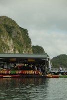 A floating structure houses numerous colorful kayaks lined up neatly in rows against a backdrop of lush green cliffs. The reflection of the kayaks and the structure is visible in the calm water. People are seen near the kayaks, suggesting an outdoor activity or rental area.