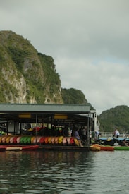 A floating structure houses numerous colorful kayaks lined up neatly in rows against a backdrop of lush green cliffs. The reflection of the kayaks and the structure is visible in the calm water. People are seen near the kayaks, suggesting an outdoor activity or rental area.