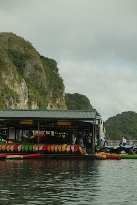 A floating structure houses numerous colorful kayaks lined up neatly in rows against a backdrop of lush green cliffs. The reflection of the kayaks and the structure is visible in the calm water. People are seen near the kayaks, suggesting an outdoor activity or rental area.