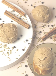 Three scoops of brown ice cream are placed on white and gold plates, surrounded by cinnamon sticks, fennel seeds, and black peppercorns on a light background.