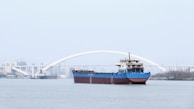A large cargo ship with a blue hull is navigating a wide river. In the background, there is a white arched bridge with the city skyline faintly visible beyond it. Another smaller ship is seen further back on the left, heading in the opposite direction.
