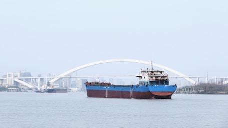 A large cargo ship with a blue hull is navigating a wide river. In the background, there is a white arched bridge with the city skyline faintly visible beyond it. Another smaller ship is seen further back on the left, heading in the opposite direction.