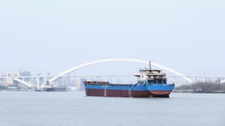 A large cargo ship with a blue hull is navigating a wide river. In the background, there is a white arched bridge with the city skyline faintly visible beyond it. Another smaller ship is seen further back on the left, heading in the opposite direction.
