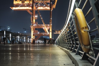 A nighttime urban scene featuring a well-lit, large industrial crane structure with a metallic walkway. Several people are walking along the path, which is bordered by a modern railing with a lifebuoy attached. The atmosphere is illuminated by a series of lights, with city buildings and cranes visible in the distant background.