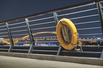 A yellow life buoy is attached to the metal railing beside a walkway. In the background, a brightly illuminated bridge spans across the water, and city lights are visible, reflecting on the water surface during nighttime.