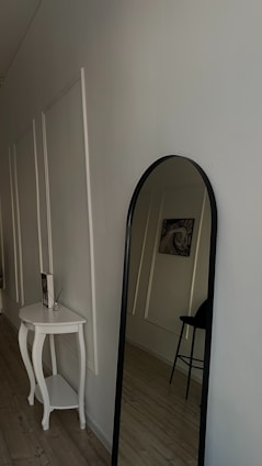 A minimalistic interior hallway features a modern, white decorative table with curved legs positioned against a wall with panel detailing. A tall, black-framed mirror is leaning against the wall next to the table. A simple artwork depicting an abstract eye design is hung on the wall. The floor is covered in light wood planks and a black stool is partially visible in the background, reflecting a clean and uncluttered atmosphere.