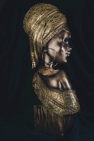 A detailed bronze sculpture of a woman adorned with intricate patterns and textures. The woman wears a headdress and large hoop earrings, exuding a sense of cultural richness and heritage. The lighting highlights the metallic sheen of the sculpture, emphasizing its artistic craftsmanship.