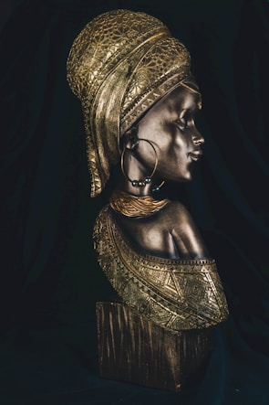 A detailed bronze sculpture of a woman adorned with intricate patterns and textures. The woman wears a headdress and large hoop earrings, exuding a sense of cultural richness and heritage. The lighting highlights the metallic sheen of the sculpture, emphasizing its artistic craftsmanship.