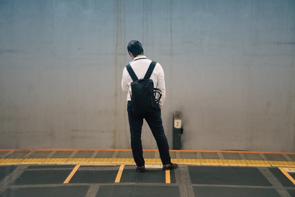 A person wearing a white shirt, black pants, and black backpack stands alone on a platform, facing away from the camera and looking downward. The platform has a yellow and black safety strip along the edge, and the backdrop is a plain gray wall.