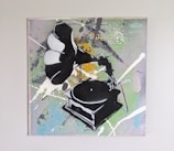 A piece of graffiti-style art featuring a phonograph with paint splatters surrounding it. The background displays a mix of colors including green, white, yellow, and some blue tones. The phonograph is depicted in a stencil-like manner, predominantly in black and white.