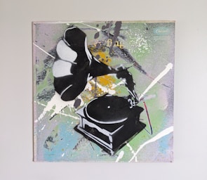 A piece of graffiti-style art featuring a phonograph with paint splatters surrounding it. The background displays a mix of colors including green, white, yellow, and some blue tones. The phonograph is depicted in a stencil-like manner, predominantly in black and white.