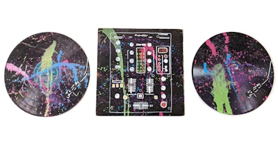 A colorful, abstract depiction of musical elements on vinyl records and a DJ mixer. The records and mixer are covered with splashes of bright neon colors including green, pink, and blue against a dark background.