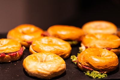 A group of bagels is arranged on a dark surface, each filled with leafy greens and slices of cured meat or similar filling. The light highlights the shiny, golden-brown crust of the bagels, creating an appetizing appearance.