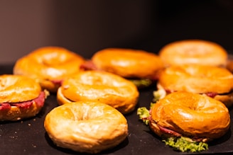 A group of bagels is arranged on a dark surface, each filled with leafy greens and slices of cured meat or similar filling. The light highlights the shiny, golden-brown crust of the bagels, creating an appetizing appearance.
