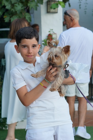 A young boy is holding a small Yorkshire Terrier dog, both dressed in white. In the background, there are people wearing casual white clothing, standing in a garden setting with green foliage and a white wall.