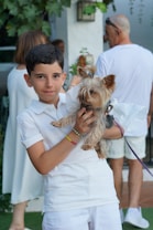 A young boy is holding a small Yorkshire Terrier dog, both dressed in white. In the background, there are people wearing casual white clothing, standing in a garden setting with green foliage and a white wall.