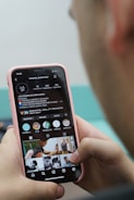 A person is holding a smartphone with their thumb on the screen, displaying an Instagram profile. The profile appears to belong to a barbershop, showcasing various images related to hairstyling. The phone case is pink, and the hands are the primary focus, with a blurred background adding depth.