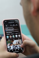 A person is holding a smartphone with their thumb on the screen, displaying an Instagram profile. The profile appears to belong to a barbershop, showcasing various images related to hairstyling. The phone case is pink, and the hands are the primary focus, with a blurred background adding depth.