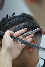 A close-up of a haircut in progress, focusing on a section of short dark hair being trimmed with scissors. A hand is steadying the hair with a comb, and a few small hair clippings are visible.