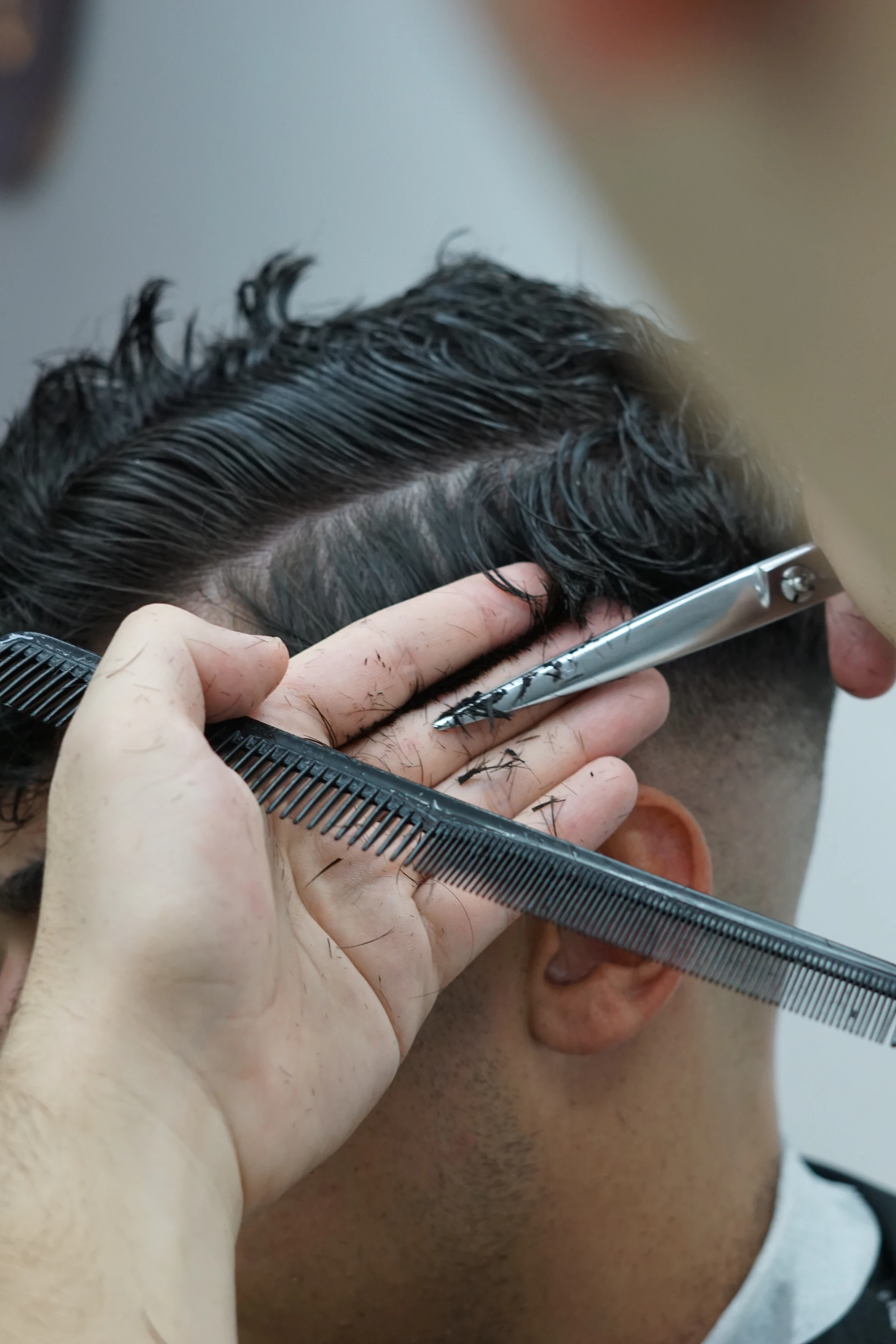 Close-up of a comb and shears cutting a section of wet dark hair held between two fingers above the ear