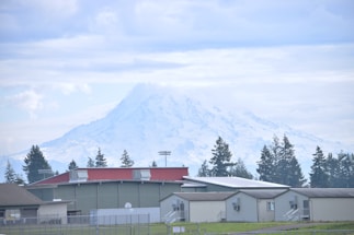 A snow-capped mountain dominates the background with large industrial buildings and a few evergreen trees in the foreground. The sky is overcast with light clouds, and the overall scene conveys a sense of calm and spaciousness.