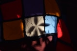 A patchwork of crocheted squares in various colors including red, orange, blue, yellow, and cream. The pattern features shadow play with a floral or star-shaped shadow cast across the squares, creating a visually striking contrast between light and darkness.