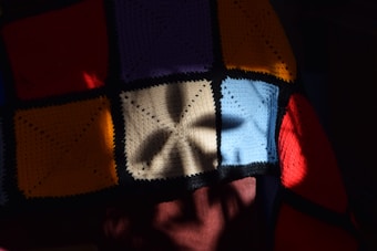 A patchwork of crocheted squares in various colors including red, orange, blue, yellow, and cream. The pattern features shadow play with a floral or star-shaped shadow cast across the squares, creating a visually striking contrast between light and darkness.