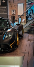 A sleek and luxurious sports car with a carbon fiber body and gold alloy wheels is displayed on a polished wooden platform. The setting includes glass barriers, informational plaques, and a staircase in the background, creating a sophisticated showroom environment.