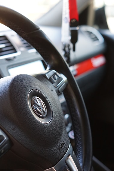 The image features a close-up view of a car's interior, specifically focusing on a Volkswagen steering wheel. Adjacent to the steering wheel, various controls and a portion of the dashboard are visible. A red and white keychain or accessory is hanging, and part of the car's air vents and infotainment system is present.