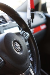 The image features a close-up view of a car's interior, specifically focusing on a Volkswagen steering wheel. Adjacent to the steering wheel, various controls and a portion of the dashboard are visible. A red and white keychain or accessory is hanging, and part of the car's air vents and infotainment system is present.